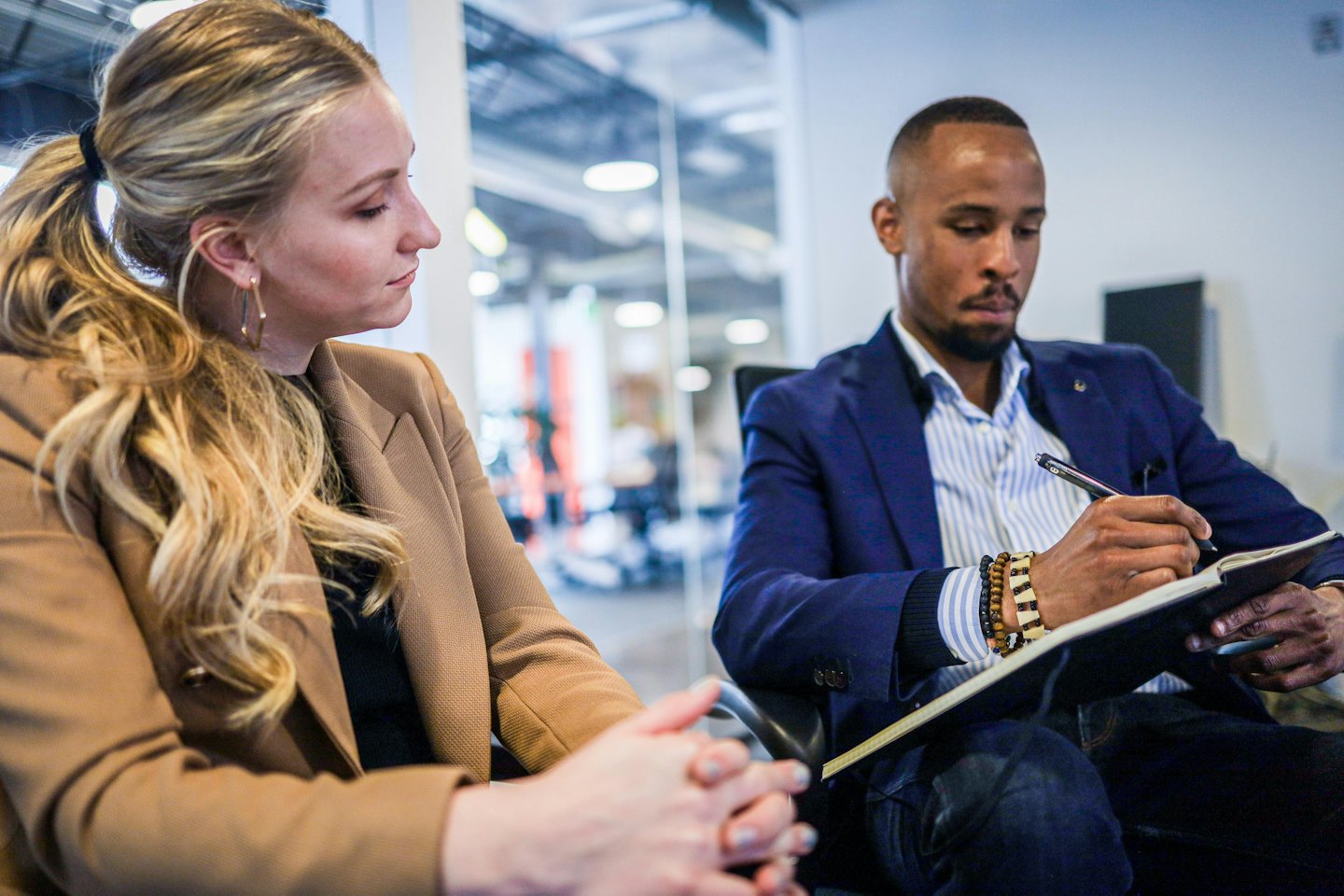 Woman in tan blazer listens to man taking notes in office.
